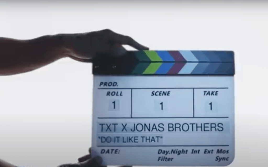 Mãos segurando uma claquete em cena do MV da música Do It Like That com Jonas Brothers e TXT