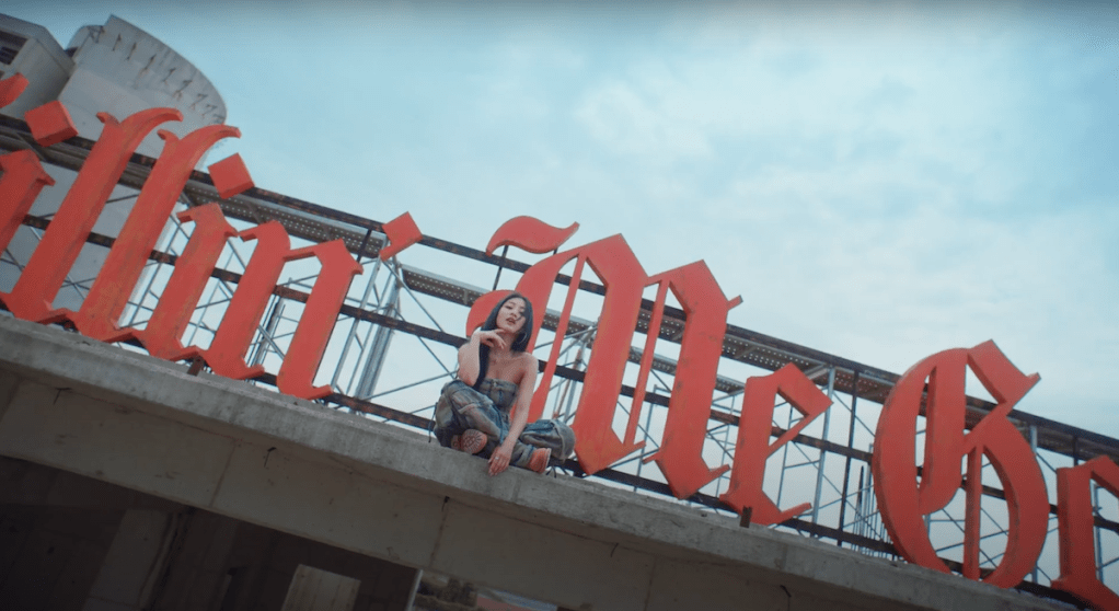 Jihyo sentada em cena do MV de Killin' Me Good do álbum ZONE com o nome da titletrack em letreiro no fundo