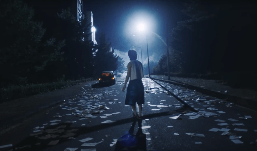 Cena do MV de Guilty com Taemin de costas para a câmera em um cenário escuro, apenas um poste iluminando vários papéis jogados no chão