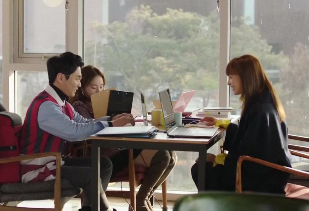 Cena de Romance is a Bonus Book com a equipe da editora de livros trabalhando em uma mesa com computadores