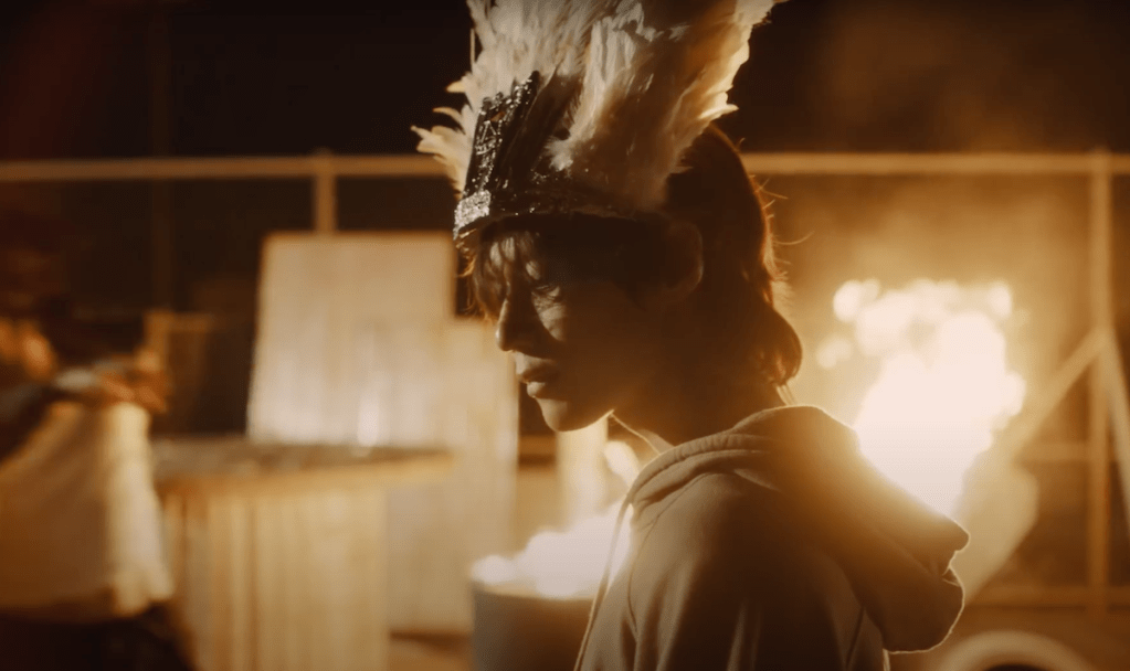 Cena do MV de Guilty com Taemin usando um cocar na cabeça e fogo no fundo do cenário