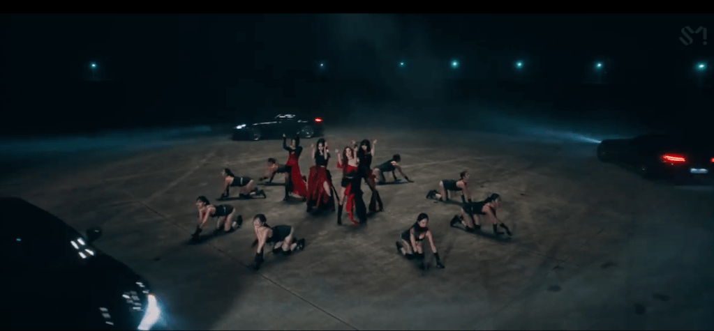 Cena do MV de "Drama", titletrack dio álbum Drama do grupo vespa com Karina, Ningning, Winter e Giselle dançando, dançarinas na volta e carros circulando 