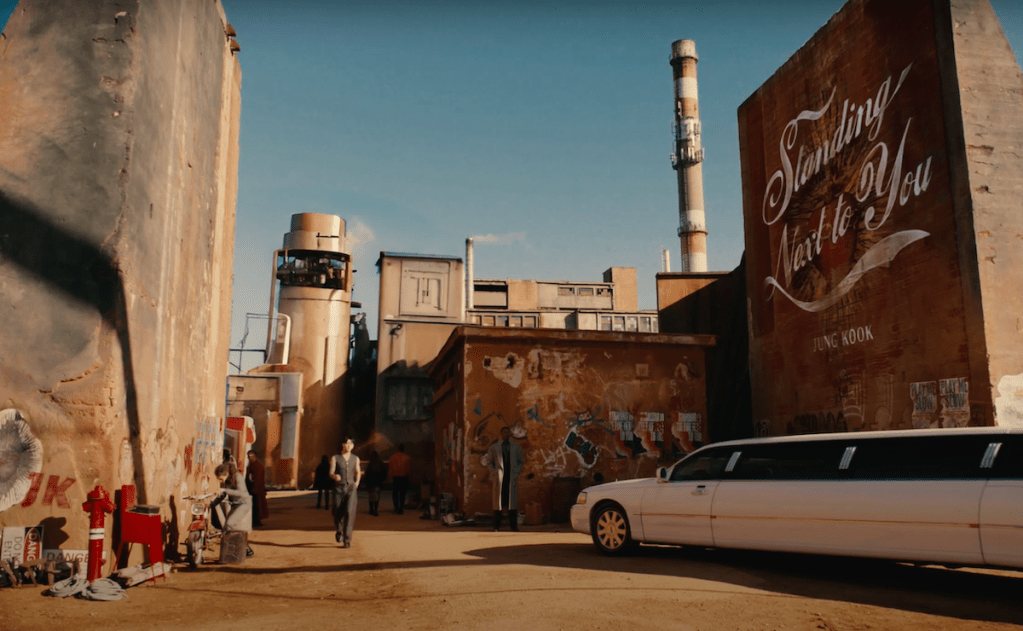 Cena do MV de "Standing Next to You" do album Golden com uma imagem de um galpão e uma limousine branca chegando