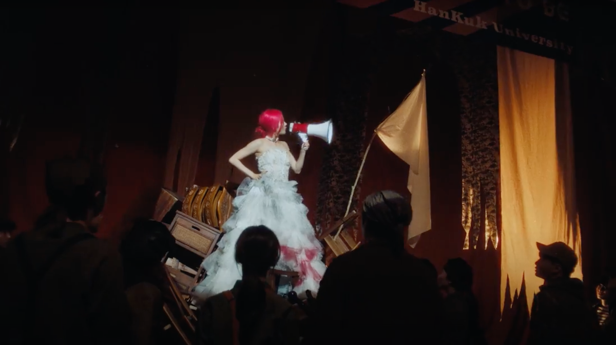 Cena do MV de "Bad News" do álbum Born to be XX do KISS OF LIFE com Belle em um palco segurando um alto-falante