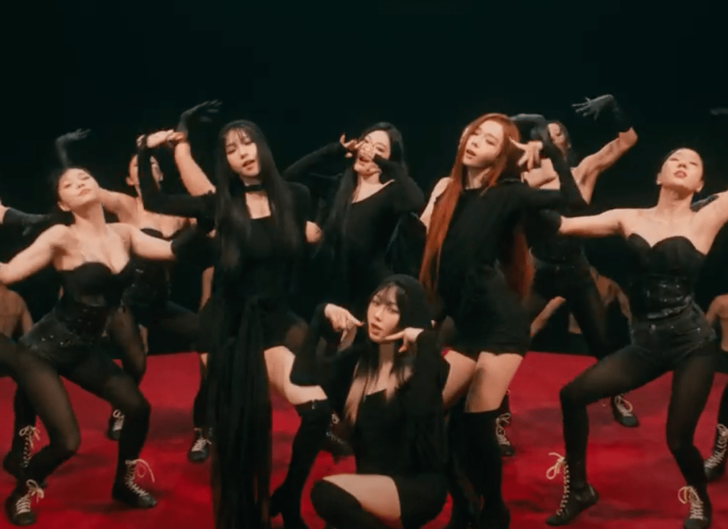 Cena do MV de "Drama", titletrack dio álbum Drama do grupo vespa com Karina, Ningning, Winter e Giselle dançando com roupas pretas em contraste com o chão vermelho