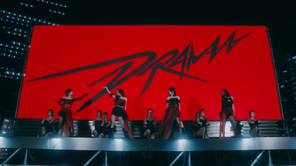 Cena do MV de "Drama", titletrack dio álbum Drama do grupo vespa com Karina, Ningning, Winter e Giselle na frente de um grande outdoor vermelho com Drama escrito em destaque