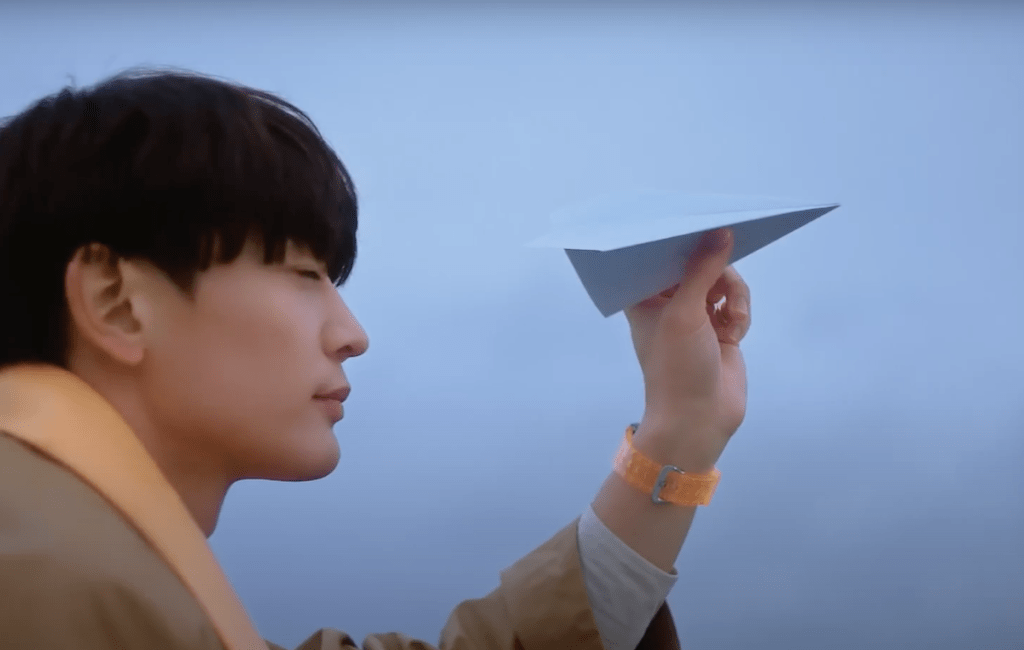 Minho (SHINee) em cena do MV de "Stay For a Night", segurando um avião de papel com os olhos fechados