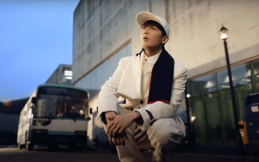 Minho (SHINee) em cena do MV de "Stay For a Night", abaixado no meio da cidade, com um ônibus ao fundo
