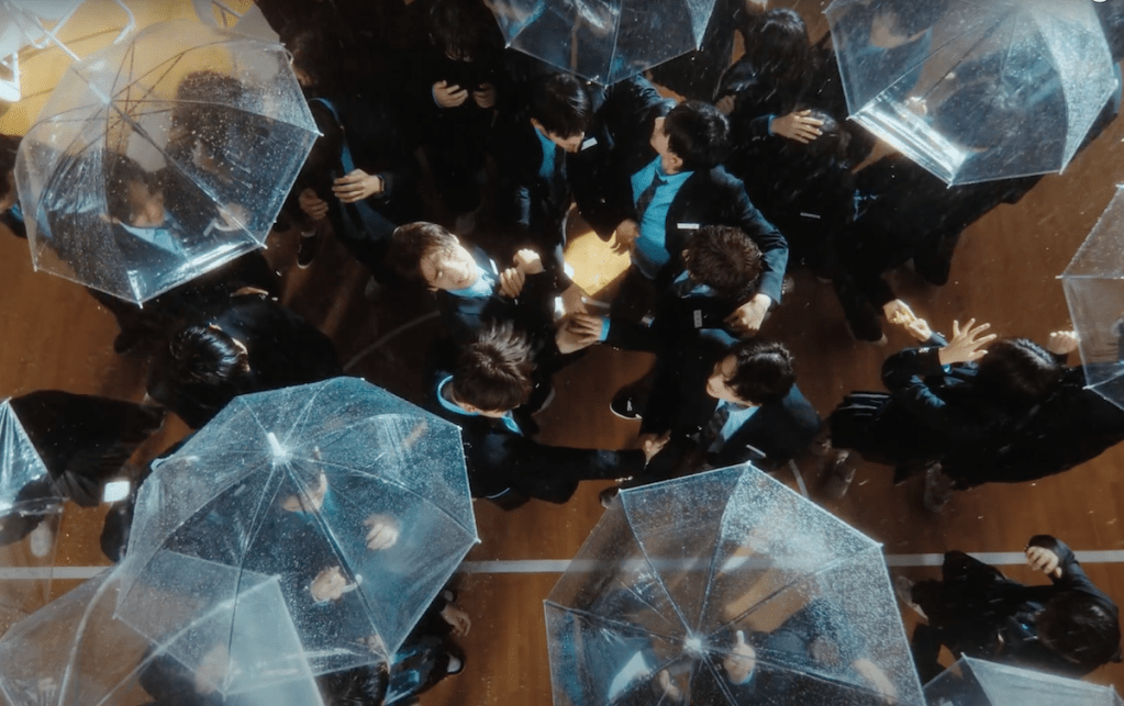 Cena do MV de "plot twist" do album Sparkling Blue com os meninos do TWS no meio de uma multidão enquanto pessoas passam com sombrinhas transparentes