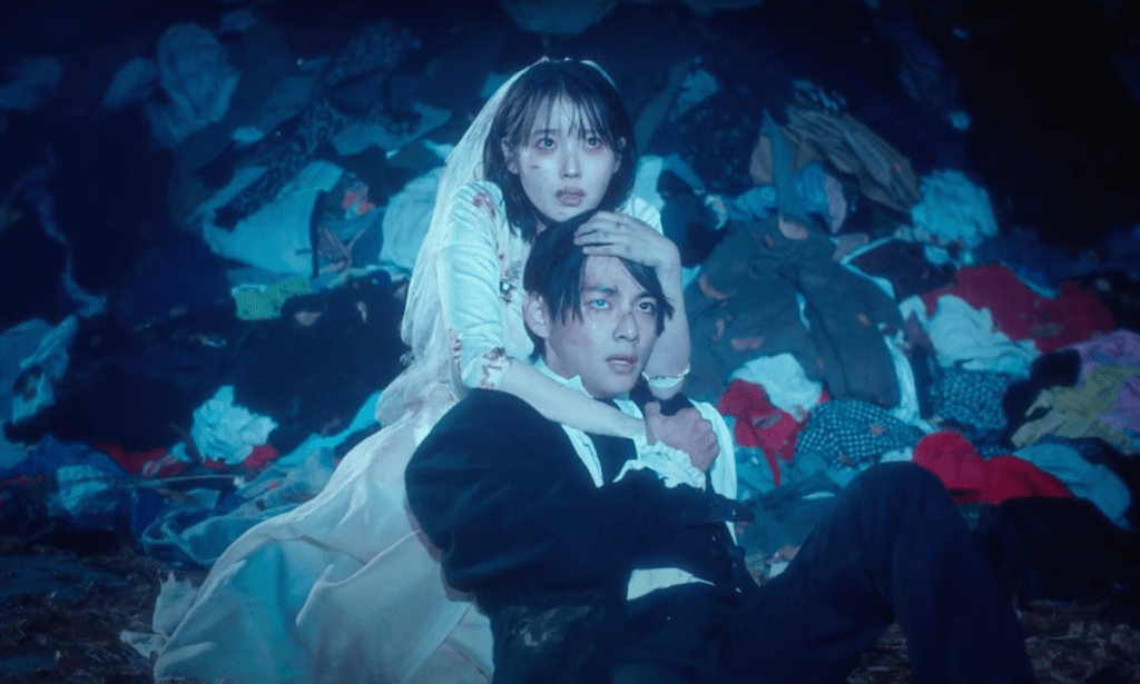 Cena do MV do single "Love wins all" com IU vestida de noiva enquanto abraça V. No fundo, uma pilha de roupas. E os dois olham para algo de forma desesperada