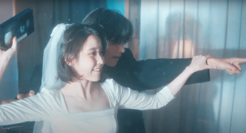 Cena do MV do single "Love wins all" com IU vestida de noiva e segurando uma fita cassete, V ao seu lado