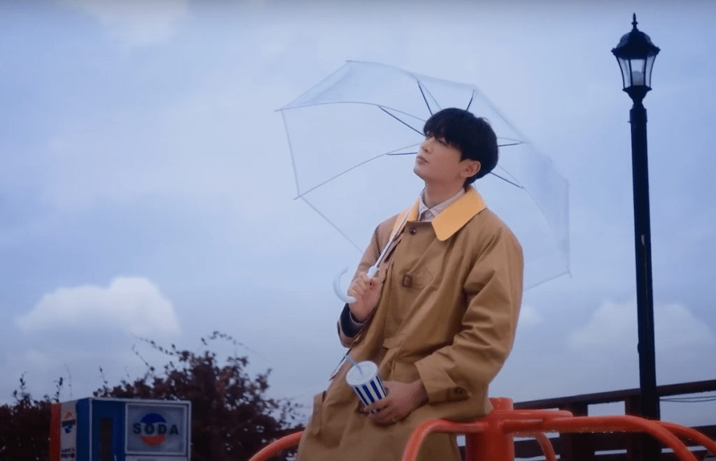 Minho (SHINee) em cena ao ar livre do MV de "Stay For a Night", segurando um guarda-chuva transparente olhando para o céu