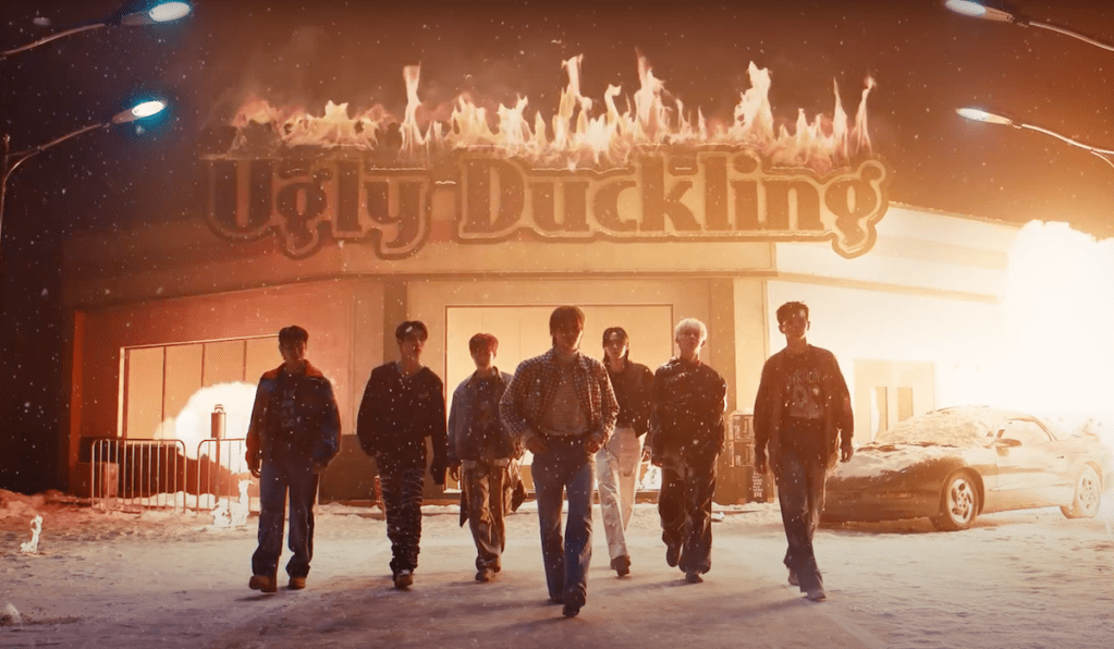 Cena do MV de "UGLY" do álbum Un: SEEN do EVNNE com os membros andando em direção à câmera, enquanto a fachada de um estabelecimento ao fundo pega fogo