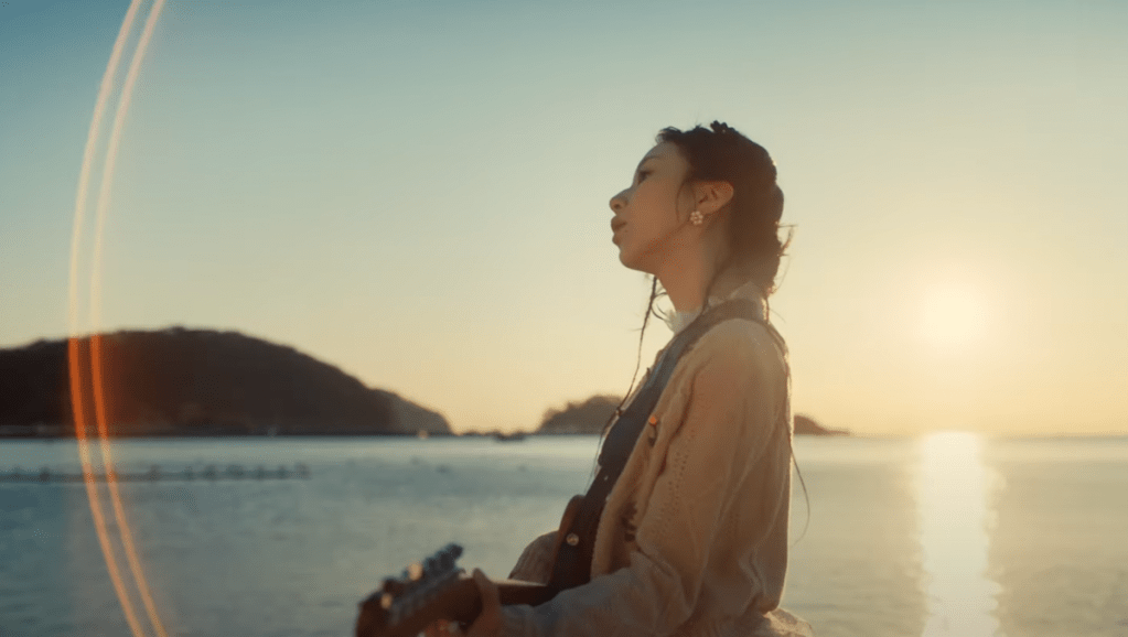 Cena do MV de "I Got You" do TWICE com Chaeyoung tocando violão, enquanto o sol de põe no mar ao fundo