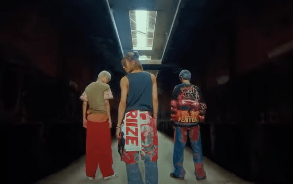 Cena com Wonbin do RIIZE de costas ao lado de dois membros no MV do single "Impossible" em uma estação de metrô
