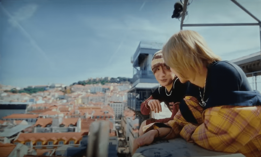 Cena do MV do single "Impossible" do RIIZE com Anton e Wonbin acompanhados pela vista para a cidade de Lisboa, em Portugal