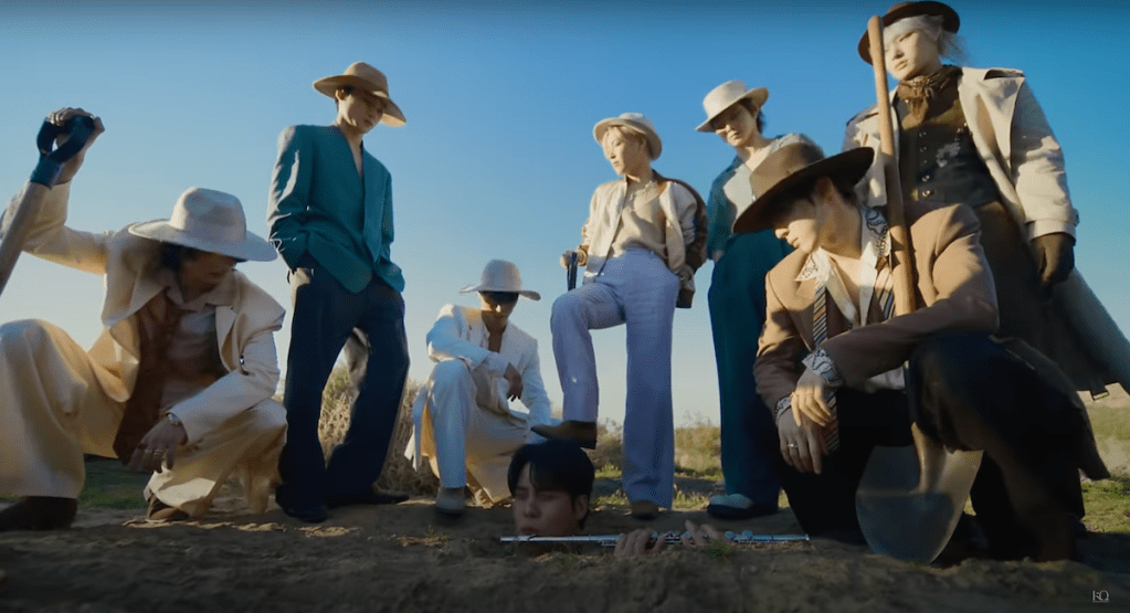 Cena do MV de "WORK" do álbum Golden Hour: Part.1 com todos os membros do ATEEZ ao ar livre, em um campo aberto e com enxadas nas mãos