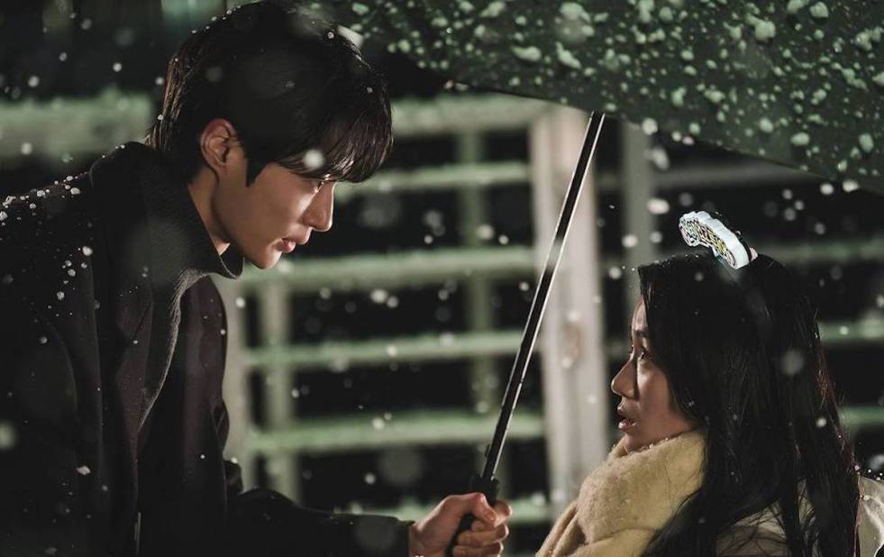Cena do K-Drama Lovely Runner com Ryu Seon-Jae (Byeon Woo-Seok) estendo um guarda-chuva para Lim Sol (Kim Hye-Yoon) em uma noite com a neve caindo