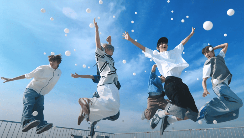 Cena do MV de "If I'm S, Can You Be My N?" do álbum SUMMER BEAT com todos os membros no ar, cercados por bolinhas brancas e o céu azul ao fundo