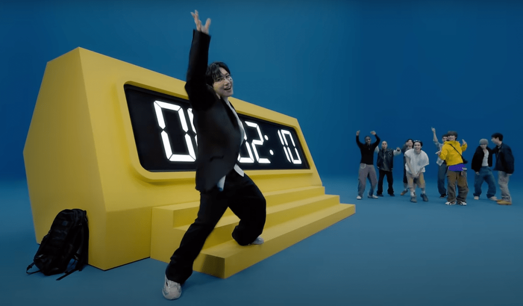 Cena do MV de "Give Me That" com os membros do WayV animados dançando perto de um grande relógio digital amarelo