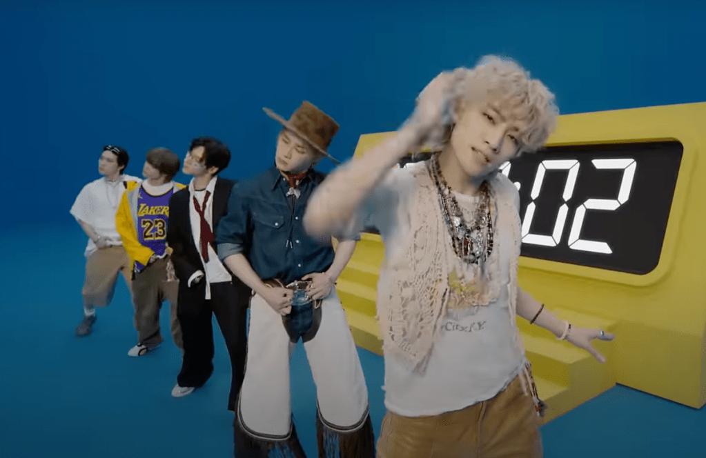 Cena do MV de "Give Me That" com os membros do WayV alinhados em fila na frente de um grande relógio digital amarelo