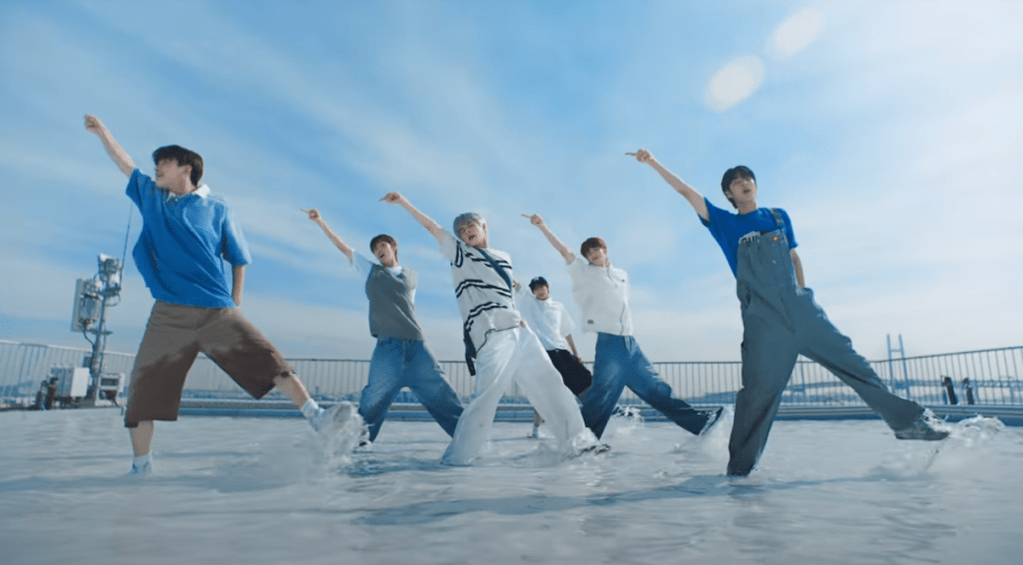 Cena do MV de "If I'm S, Can You Be My N?" do álbum SUMMER BEAT com todos os membros dançando ao ar livre em um piso coberto por água