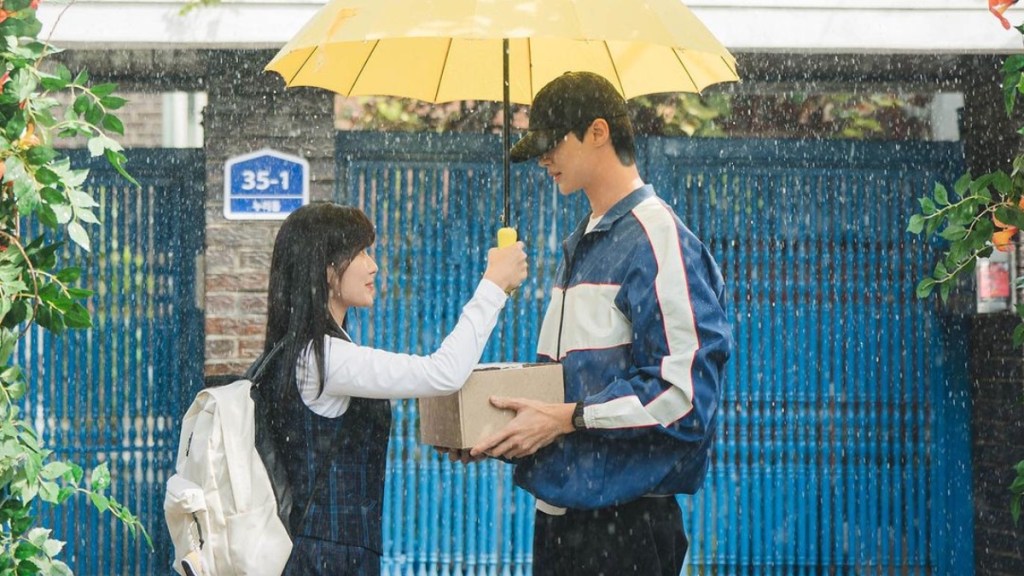 Cena do K-Drama Lovely Runner com Im Sol estendendo uma sombrinha amarela em direção ao Sun Jae na frente do portão da sua casa em um dia chuvoso