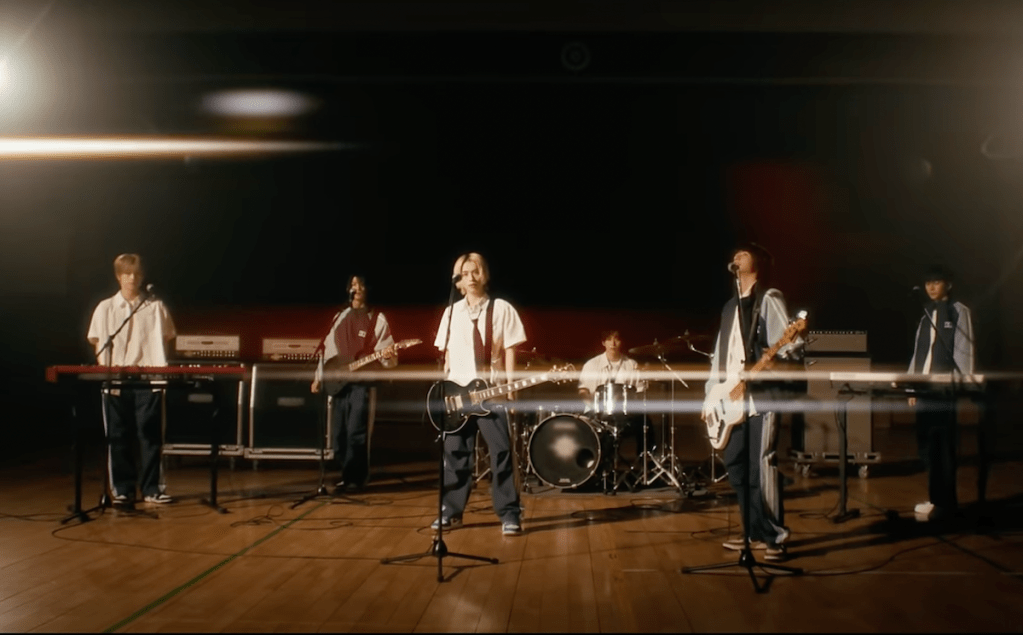 Cena do MV de Boy Comics com todos os membros da banda Xdinary Heroes e seus instrumentos musicais em um ambiente com pouca iluminação