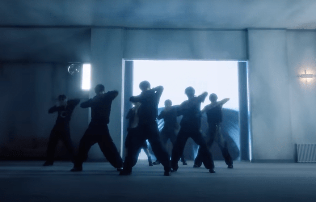 Cena do MV de "XO (Only If You Say Yes)" do álbum ROMANCE : UNTOLD do ENHYPEN com cena de coreografia com todos os membros: Sunghoon, Ni-ki, Heeseung, Jay, Sunno, Jake e Jungwon em um cenário escuro, apenas as silhuetas aparecem