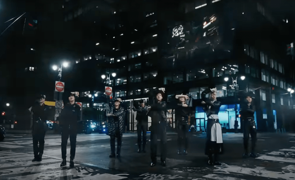 Cena do MV de "Chk Chk Boom" do álbum ATE com todos os membros do Stray Kids dançando em pose para o refrão, cercados pelas luzes noturnas dos prédios ao redor
