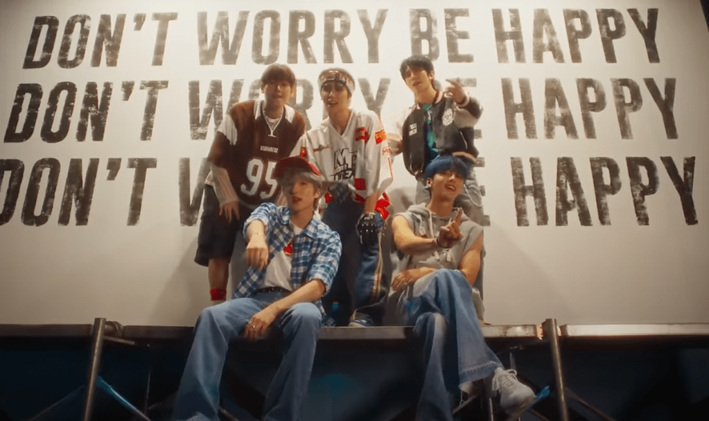 Cena do MV de "Don't Worry, Be Happy" do SF9 com o álbum Fantasy, mostrando Cha Ni, Hwi Young, In Seong, Yoo Tae Yang e Young Bin na frente de um outdoor com o nome da música