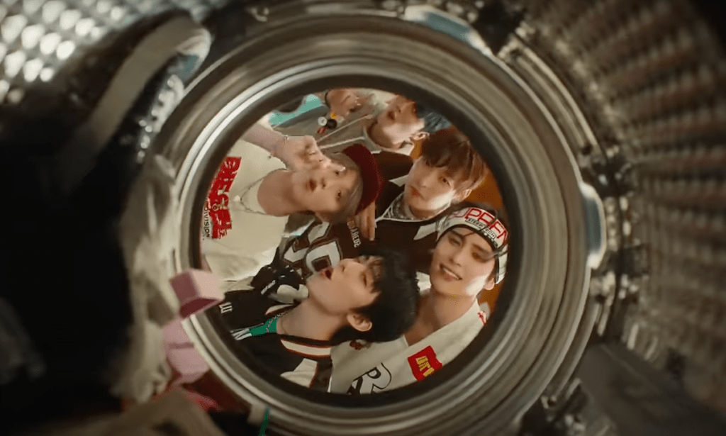 Cena do MV de "Don't Worry, Be Happy" do SF9 com o álbum Fantasy, mostrando Cha Ni, Hwi Young, In Seong, Yoo Tae Yang e Young Bin em um ângulo de dentro de uma máquina de lavar