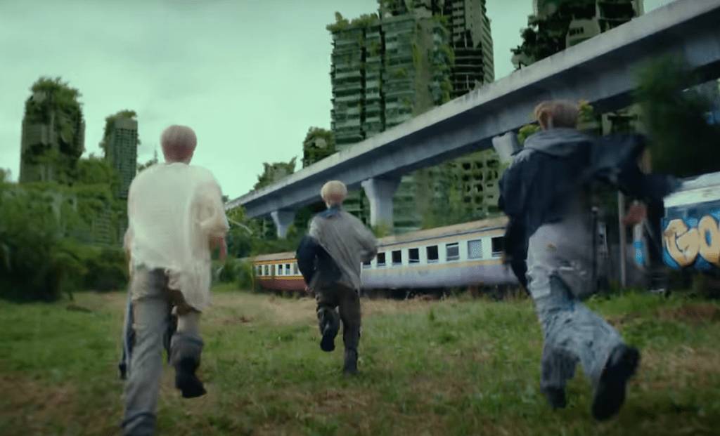 Cena do MV de "Rains in Heaven", single em inglês do NCT Dream com alguns dos membros correndo em um cenário com prédios cobertos por vegetação