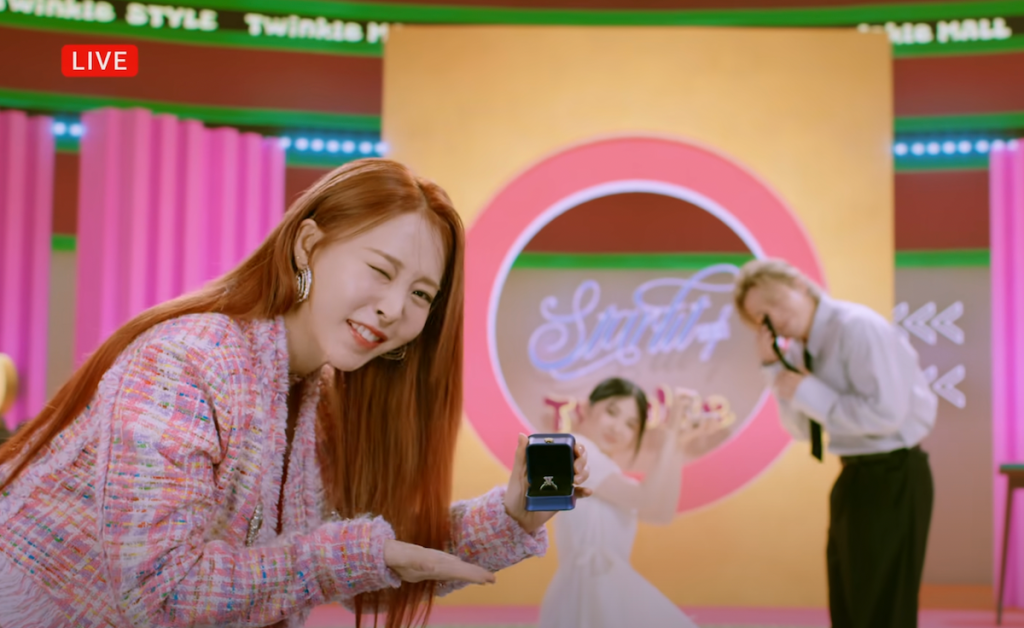 Cena do MV de "Is This Love?" do álbum Starlit of Twinkle com Moon Byul segurando uma caixinha com um anel enquanto pisca para a câmera 