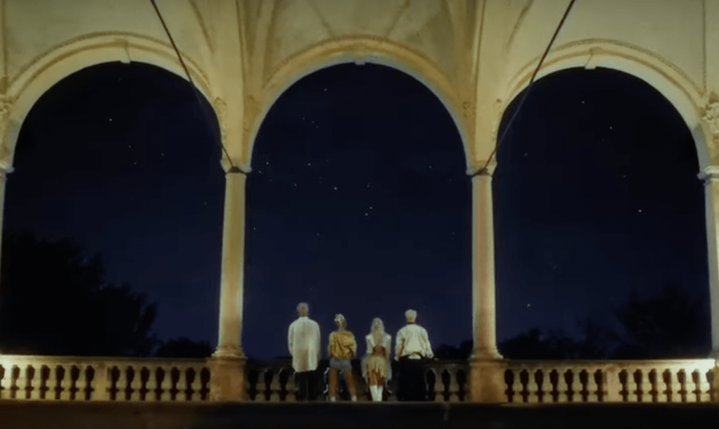 Cena do MV de "Tell My Momma" do KARD com os quatro membros de costas para a câmera, contemplando o céu estrelado