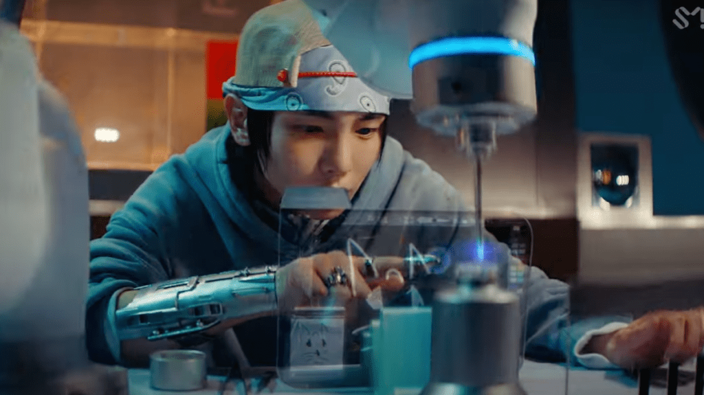 Cena do MV de "Pleasure Shop" com Key escrevendo algo em uma tela futurista em uma sala que parece um laboratório