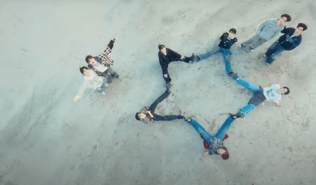 Cena do MV de "BLUE" do álbum Blue Paradise do ZB1. Os meninos estão sentados na areia formando o desenho de uma estrela com as pernas