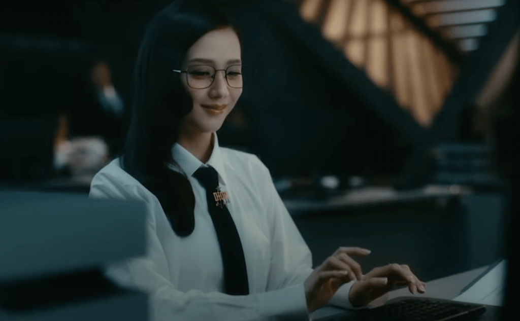 Cena do MV de earthquake do álbum AMORTAGE da Jisoo. Ela veste roupas de escritório e sorri quando olha para o seu celular