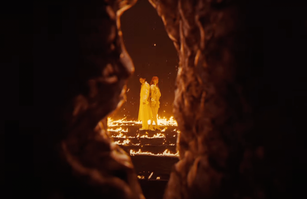 Cena do MV de "ReawakeR" da collab de LiSA com Felix do Stray Kids. Os dois podem ser vistos através de uma fenda, cercados por um cenário em chamas