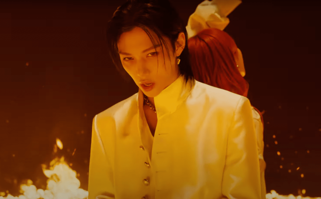 Cena do MV de "ReawakeR" da collab de LiSA com Felix do Stray Kids. Foco no Felix, enquanto ele olha para a câmera em um cenário em chamas