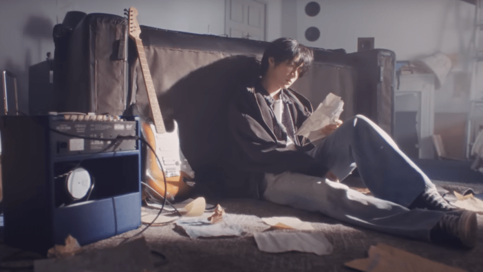 Cena do MV de Panic com o debut solo de Beomgyu do TXT, que aparece na imagem sentado no chão e cercado por uma guitarra, equipamentos musicais e rascunhos de letras