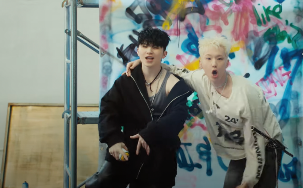 Cena do MV de 96eurs do álbum BEAM, marcando o debut da unit HxW com Hoshi e Woozi do SEVENTEEN. Na imagem, os dois estão lado a lado com uma pintura colorida de spray no fundo