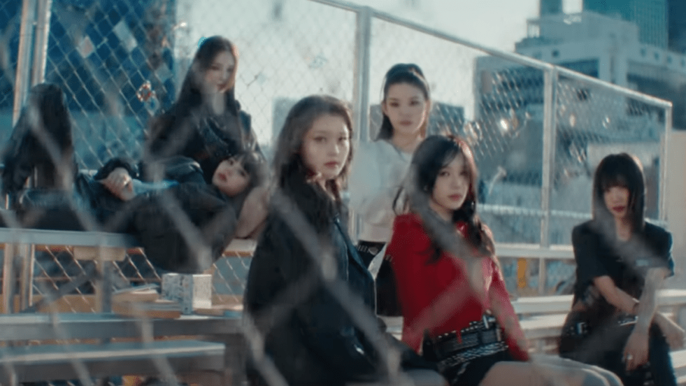 Cena do MV de "SIGN" com as integrantes do izna olhando para a câmera através de uma cerca