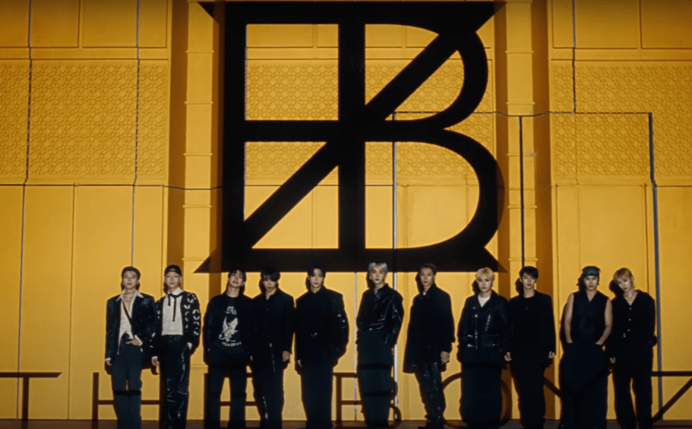 Cena do MV de "VVV" do novo álbum Unexpected do THE BOYZ com todos os membros, lado a lado, em um fundo amarelo com as iniciais do grupo