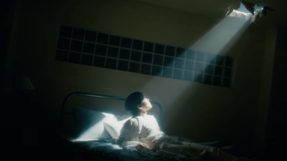 Cena simbólica do MV de Maybe Tomorrow do DAY6 em um quarto escuro, com uma pequena fresta de luz entrando