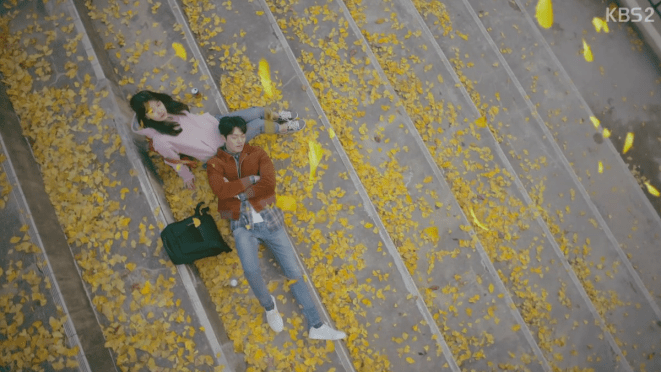 Cena do K-Drama Uncontrollably Fond em uma escada coberta por flores e folhas amarelas onde Kim Woo-Bin (Shin Joon-Young) está deitado no colo de Bae Suzy (No Eul)