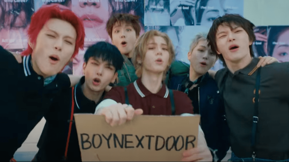 Cena do MV de "I Feel Good". do álbum No Genre do BOYNEXTDOOR com todos os membros reunidos e olhando para a câmera, enquanto seguram uma placa com o nome do grupo