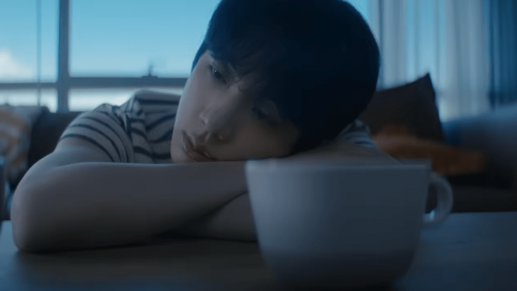 Cena do MV de "Don't Say You Love Me" do álbum Echo com Jin (BTS) pensativo, apoiado em uma mesa com uma xícara de café