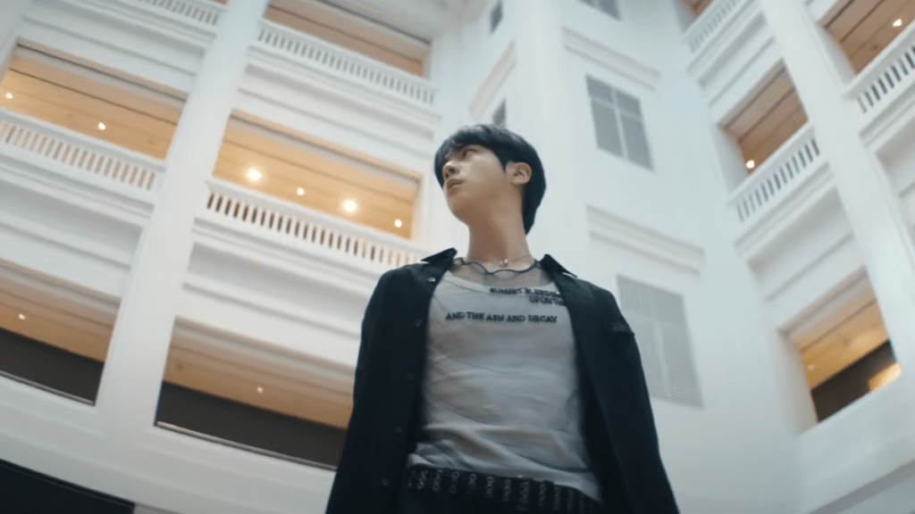 Cena do MV de "Don't Say You Love Me" do álbum Echo com Jin (BTS) olhando à sua volta e um prédio claro iluminado ao fundo