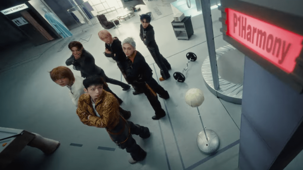 Cena do MV de "DUH!" do P1Harmony com todos os membros de braços cruzados e olhando para uma placa vermelha com o nome do grupo no canto superior direito da tela